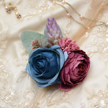 A decorative hair clip floral arrangement featuring a large blue rose, a deep burgundy ranunculus, pink chrysanthemums, and muted green leaves, styled on a cream lace bridal fabric background with a delicate necklace.