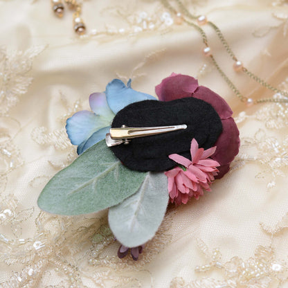 A decorative hair clip floral arrangement featuring a large blue rose, a deep burgundy ranunculus, pink chrysanthemums, and muted green leaves. Placed upside down to show a felt base and metal alligator style clip styled on a cream lace bridal fabric background with a delicate necklace