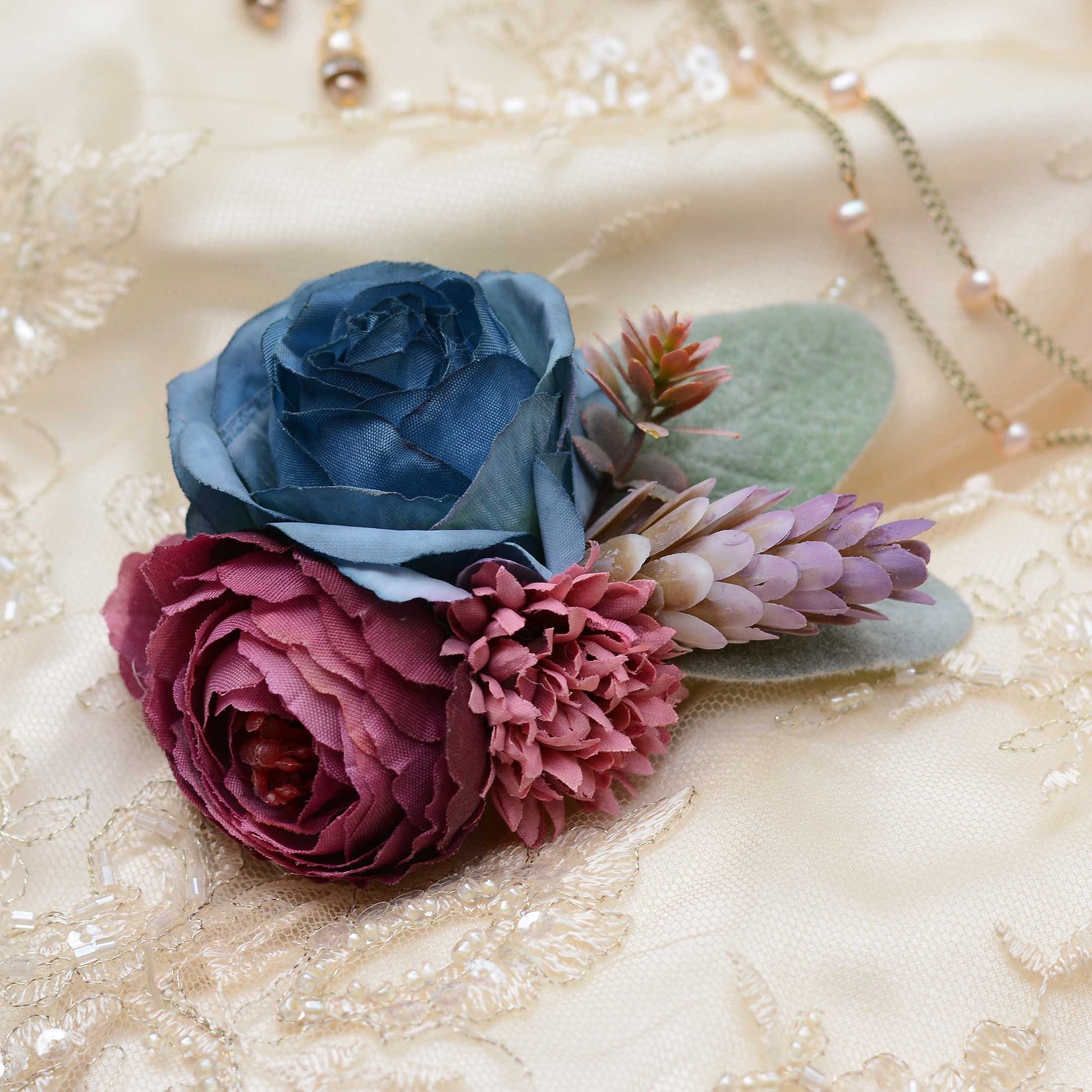 A decorative hair clip floral arrangement featuring a large blue rose, a deep burgundy ranunculus, pink chrysanthemums, and muted green leaves, styled on a cream lace bridal fabric background with a delicate necklace.