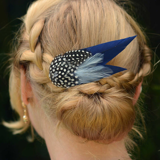 Close up of blonde braided hair in a low bun with a large navy blue feather hair clip with spotted feathers.