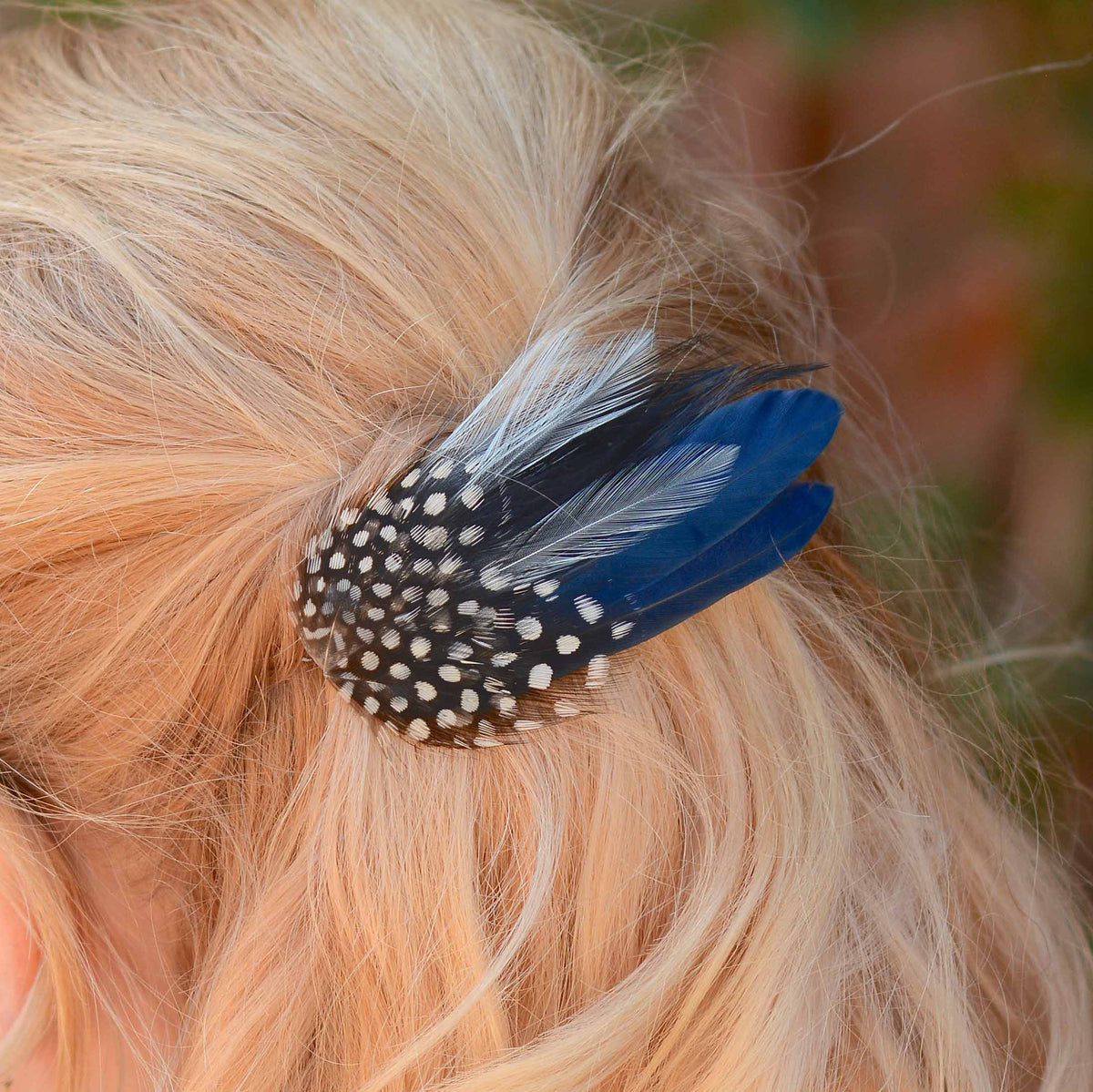 A close-up of a blonde person's hair secured with a decorative feather hair clip featuring a black-and-white spotted base and navy blue feathers.