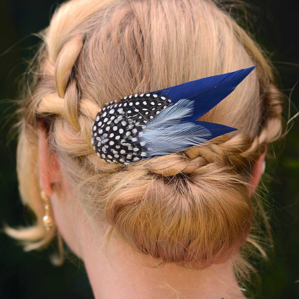 Close up of blonde braided hair in a low bun with a large navy blue feather hair clip with spotted feathers.