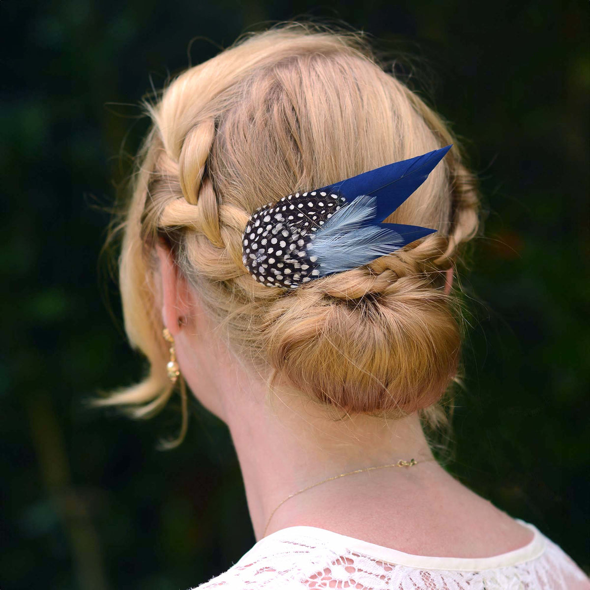 woman with blonde braided hair in a low bun with a large navy blue feather hair clip with spotted feathers.