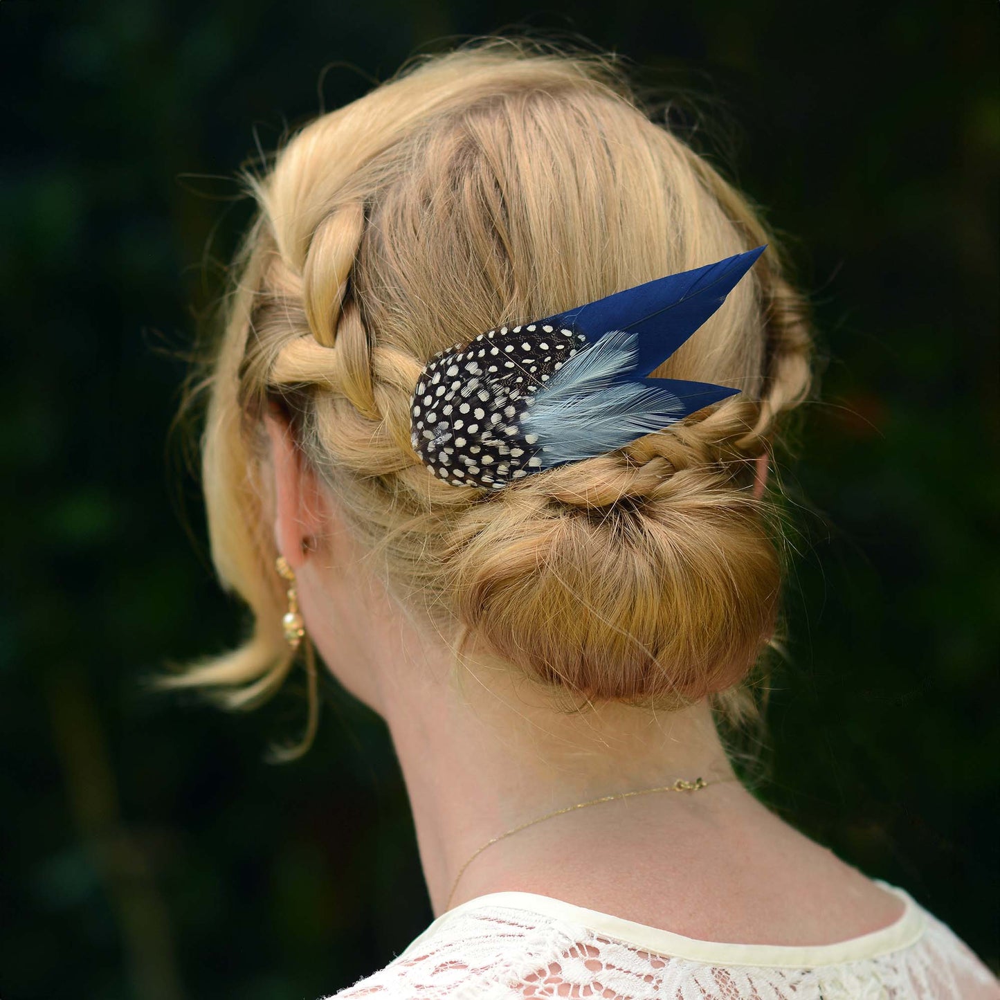 woman with blonde braided hair in a low bun with a large navy blue feather hair clip with spotted feathers.