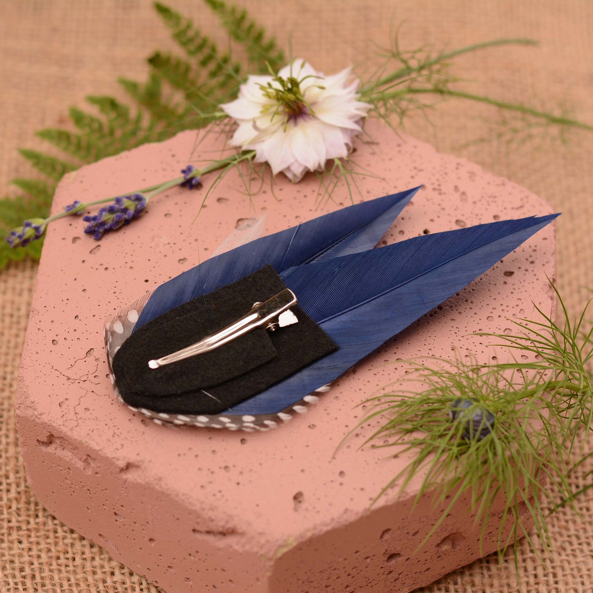 Navy blue feather hair clip with polka dot spotted guinea fowl feathers on a pink stone surrounded by greenery