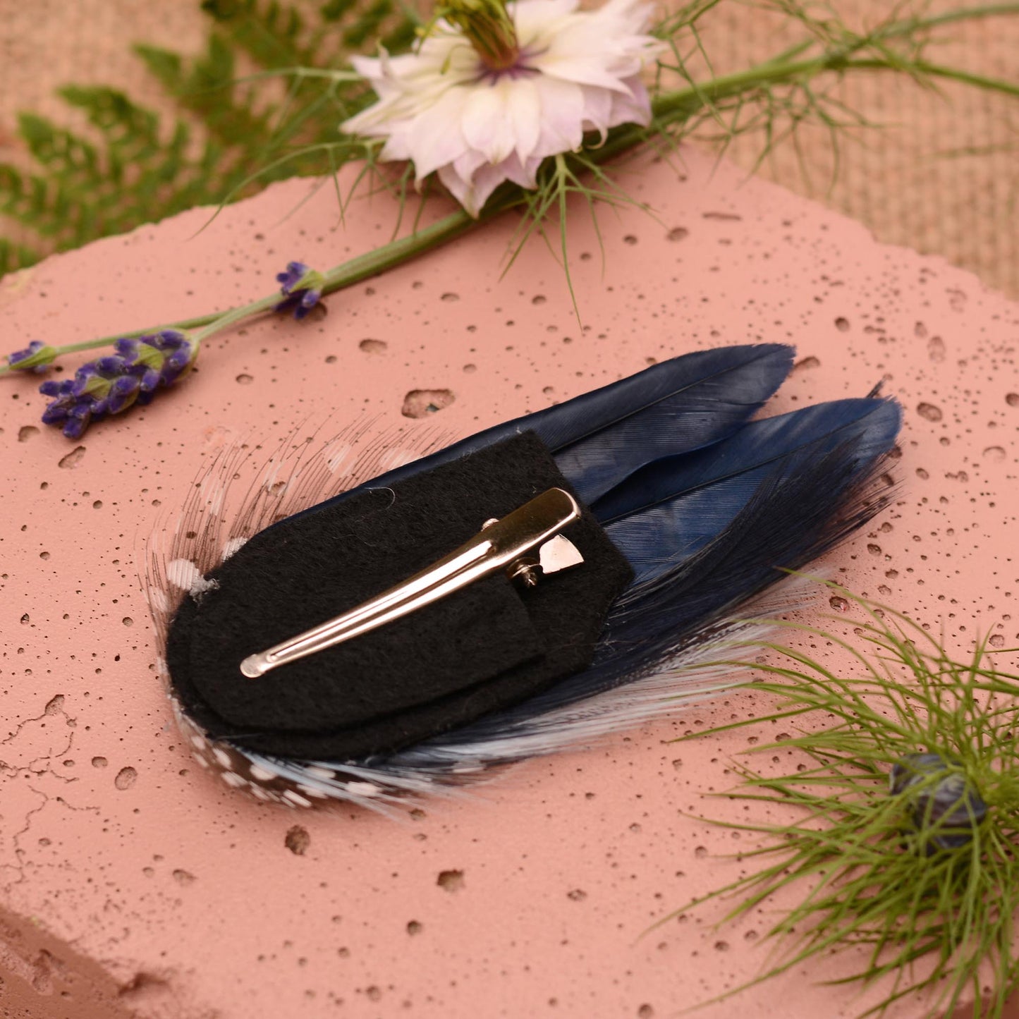 Navy blue feather hair clip with polka dot spotted guinea fowl feathers on a pink stone surrounded by greenery