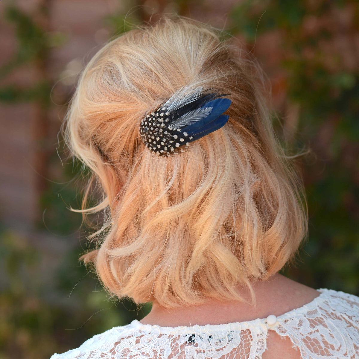 A blonde woman shown from the back wearing a feather hair clip with black and white polka dots and navy blue feathers, set against a blurred outdoor background.