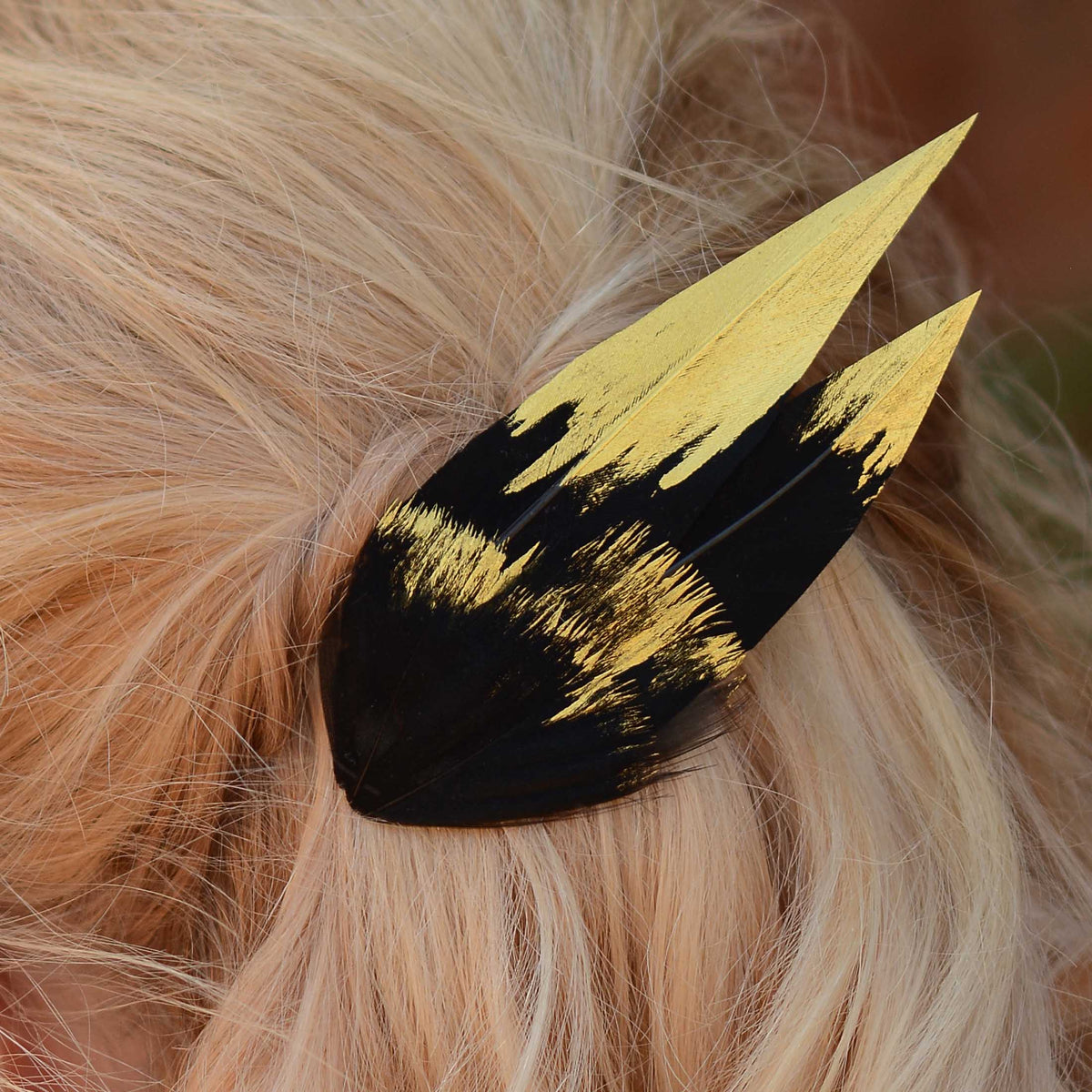 A close-up of a feather hair accessory clipped onto blonde hair. The accessory has a pointed elegant shape and features black feathers hand painted with gold tips.