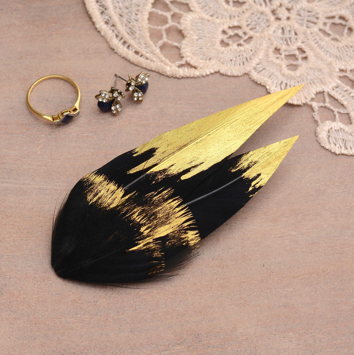 A decorative feather hair clip fascinator in black and gold, styled on a beige surface with a lace fabric, a gold ring, and small stud earrings.