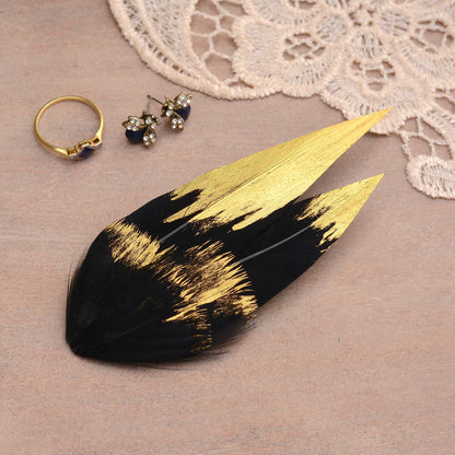 A decorative feather hair clip fascinator in black and gold, styled on a beige surface with a lace fabric, a gold ring, and small stud earrings.