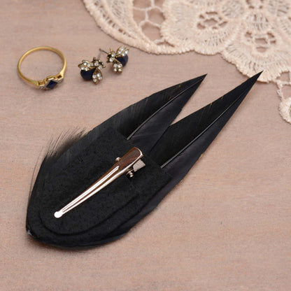 A black feather hair clip with a silver-tone metal alligator-style clip and felt base, displayed on a textured beige surface with a lace corner and small jewellery props.