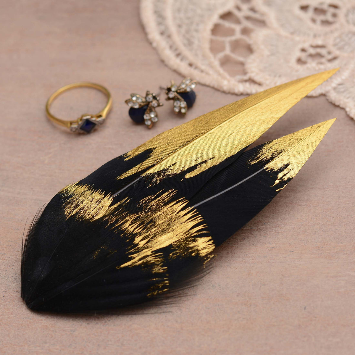 A decorative feather hair clip in black with textured gold paint strokes, displayed on a beige wood surface next to a gold ring and blue gemstone earrings.