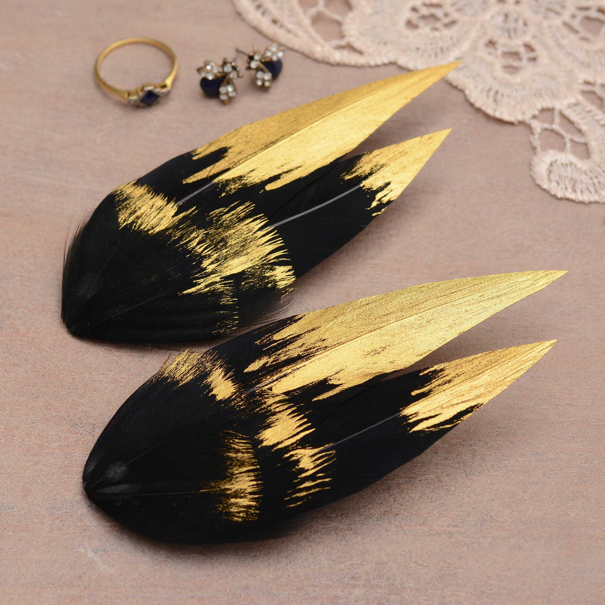 Two decorative feather hair pieces in black a with a metallic gold hand-painted brushstroke pattern, displayed on a pale wooden surface with jewellery props.