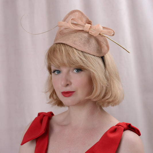 A woman with short blonde hair wearing a light nude pink side perching mother of the bride fascinator hat with a bow and gold quill feather, paired with a red dress against a soft beige curtain background.