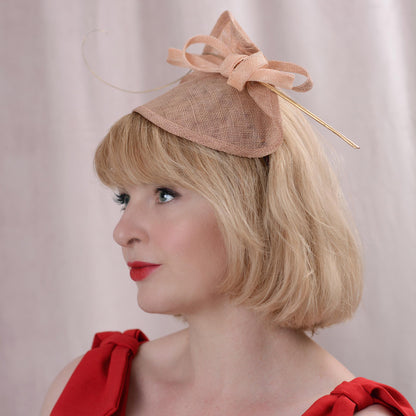 A blonde woman in profile wearing a beige fascinator hat with a large bow and a gold feather, paired with a red top against a soft beige backdrop.