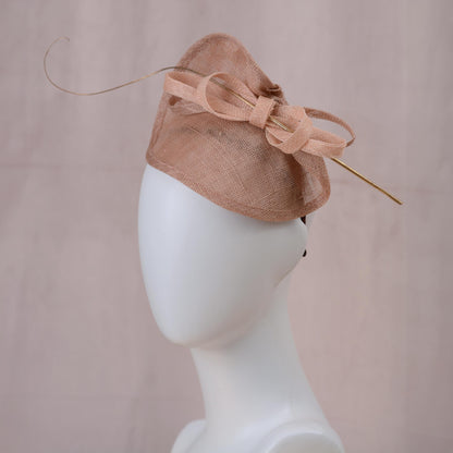 A blush pink horse racing fascinator hat with a large bow and delicate gold feather accent, displayed on a white mannequin head against a soft pink background.
