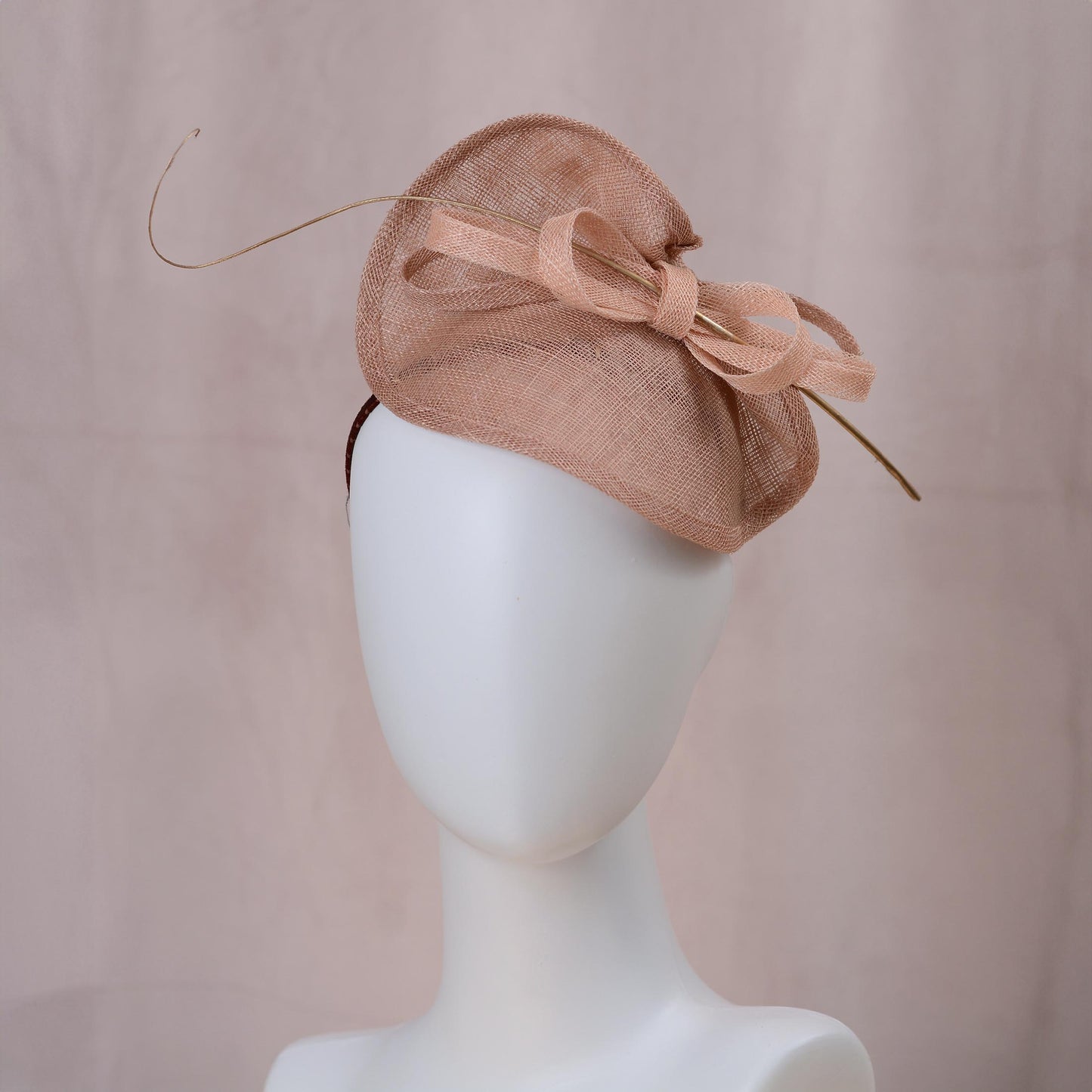 A dusty pink mother of the groom fascinator hat with a large bow and gold ostrich quill feather, displayed on a white mannequin head against a soft beige background.