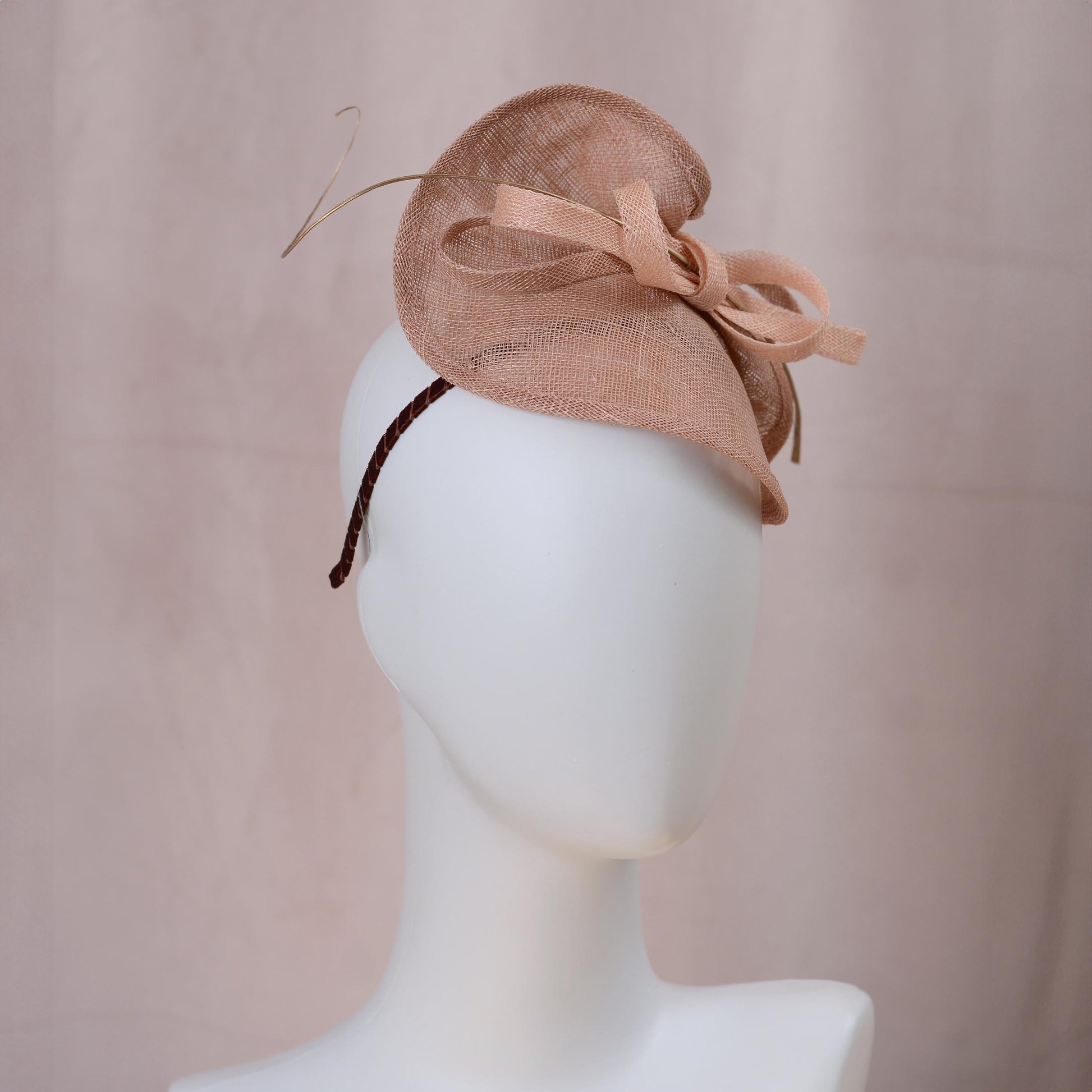 A blush pink royal ascot fascinator hat with a large bow and delicate gold feather accent, displayed on a white mannequin head against a soft pink background.