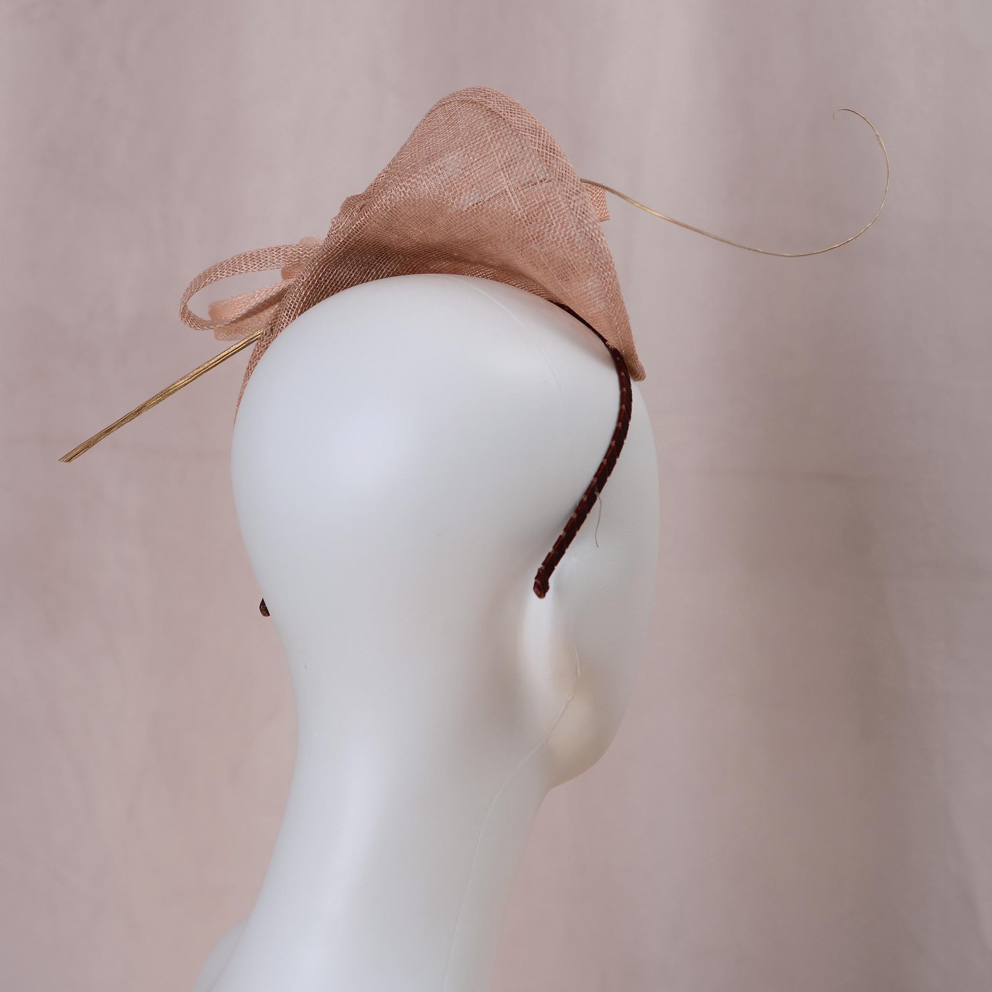 A blush pink royal ascot fascinator hat with a large bow and delicate gold feather accent, displayed on a white mannequin head against a soft pink background.