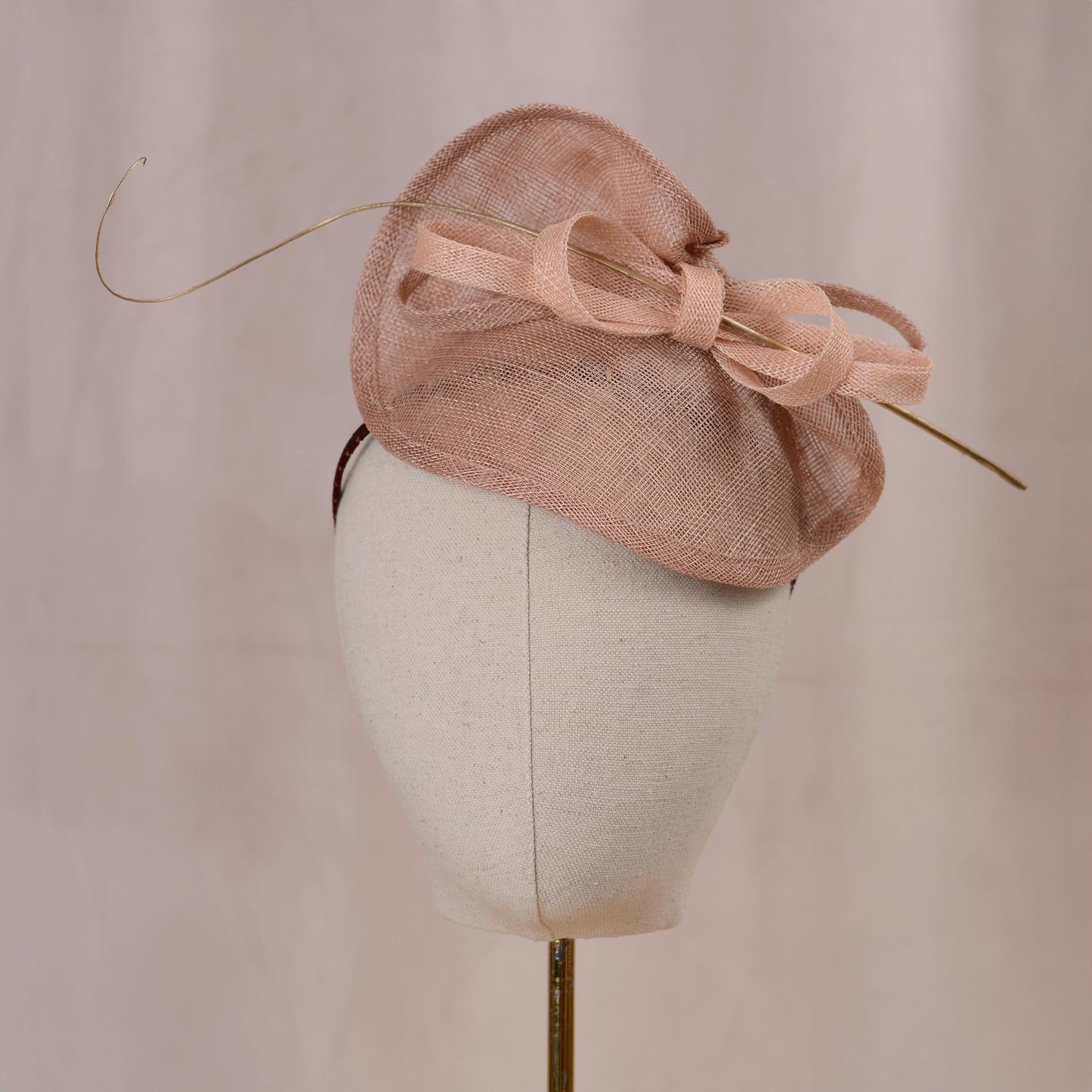 A blush pink kentucky derby fascinator hat with a large bow and delicate gold feather accent, displayed on a white mannequin head against a soft pink background.