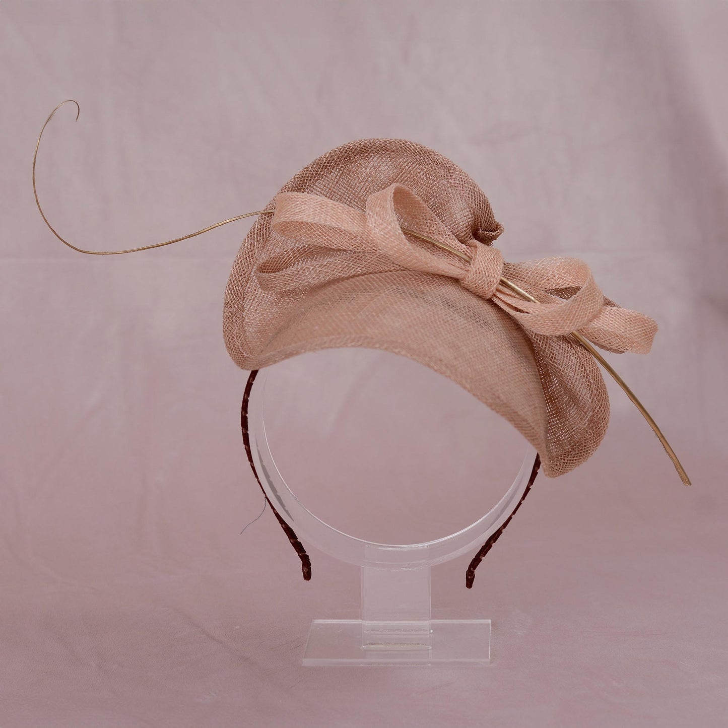 A light beige pink sinamay straw  summer wedding fascinator hat with a ribbon bow and long gold feather accent, displayed on a clear stand against a soft pink background.