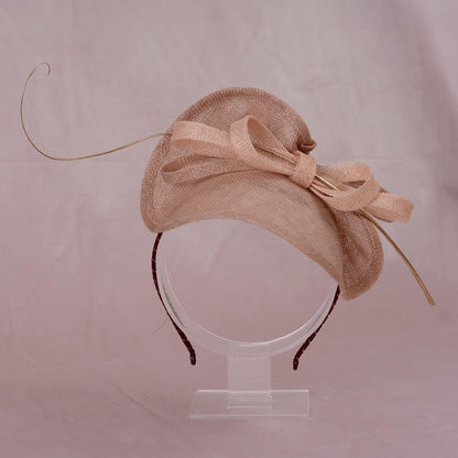 A light beige pink sinamay straw  summer wedding fascinator hat with a ribbon bow and long gold feather accent, displayed on a clear stand against a soft pink background.