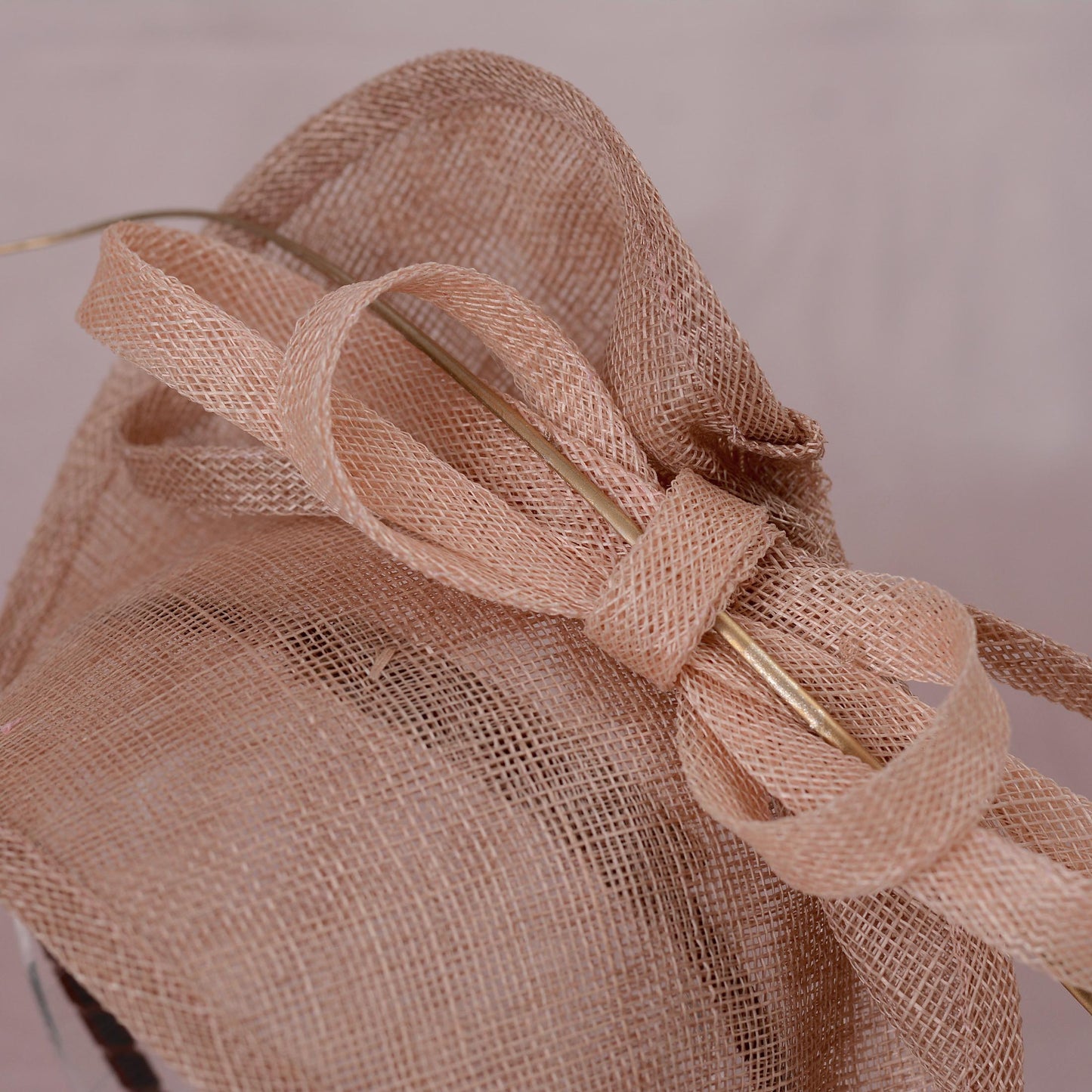 A blush pink royal garden party fascinator hat with a large bow and delicate gold feather accent, displayed on  a clear perspex hat display against a soft pink background.