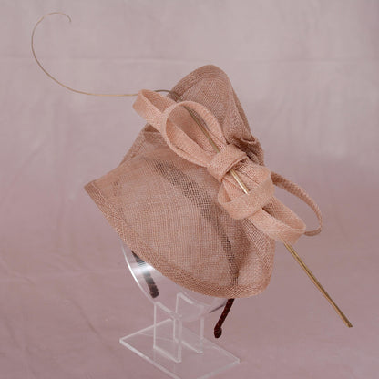 A blush pink garden party fascinator hat with a large bow and delicate gold feather accent, displayed on a clear perspex hat display against a soft pink background.