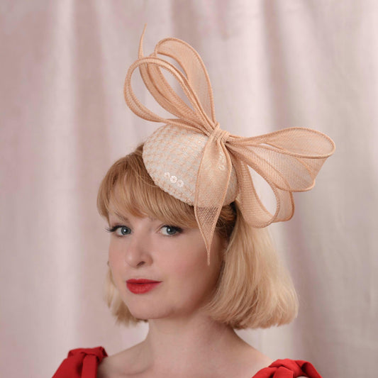 A woman with a short blonde bob wears a blush pink button wedding fascinator hat covered in an embroidered sequin fabric and trimmed with a large sinamay straw bow. She is wearing a red dress against a soft pink curtain background.