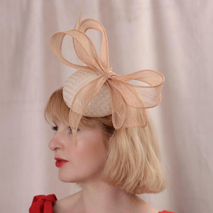 A woman with a short blonde bob wears a blush pink button royal ascot fascinator hat covered in an embroidered sequin fabric and trimmed with a large sinamay straw bow. She is wearing a red dress against a soft pink curtain background.