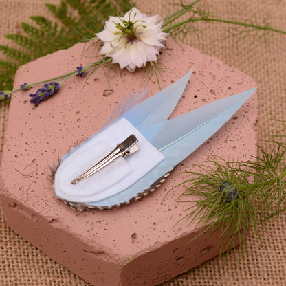 Blue feathered hair clip on a pink surface with flowers and greenery.