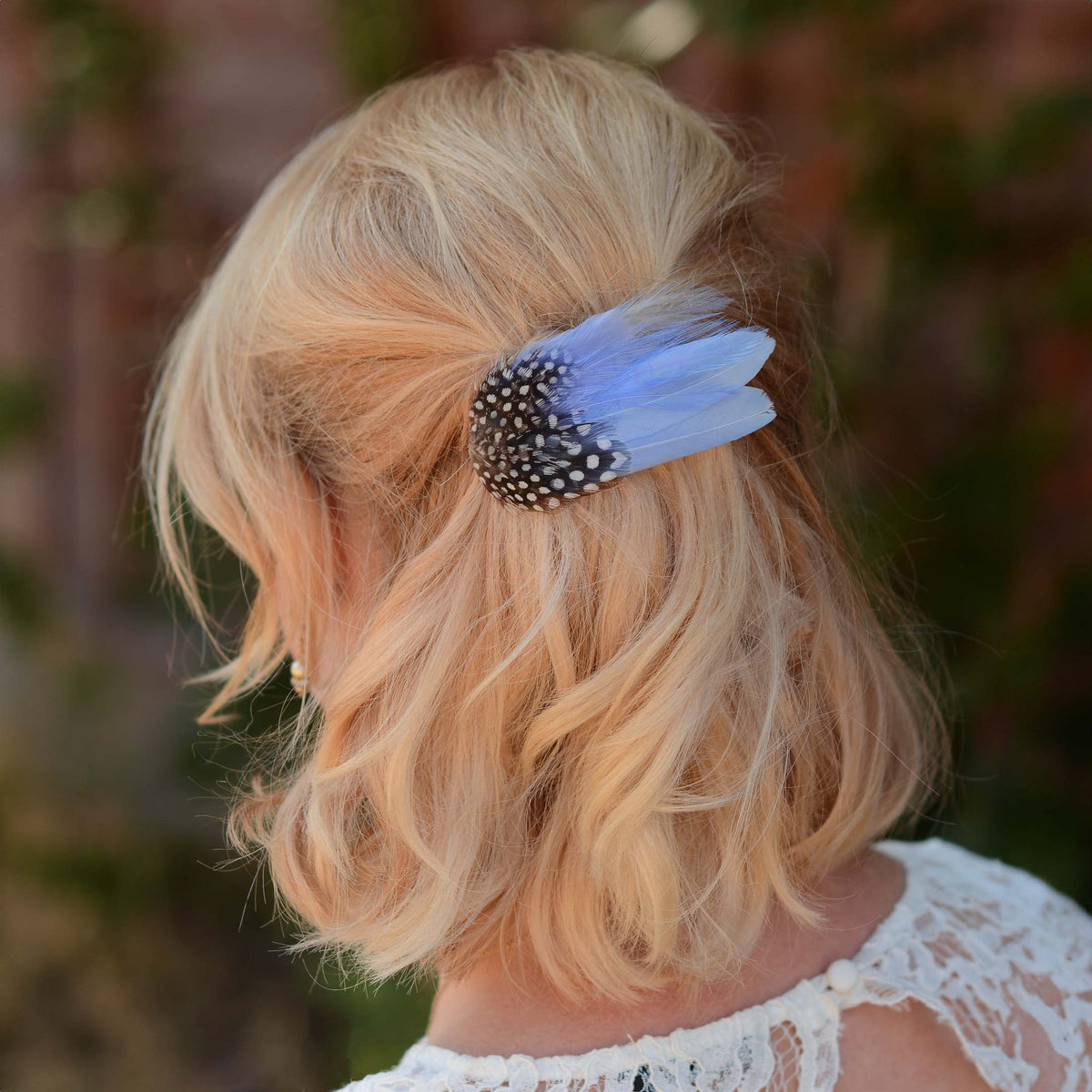A blonde woman shown from the back wearing a light blue feather hair clip with a black and white spotted feathers, set against a blurred outdoor background.