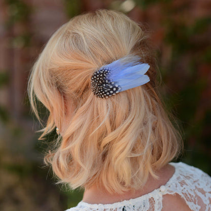 A blonde woman shown from the back wearing a light blue feather hair clip with a black and white spotted feathers, set against a blurred outdoor background.