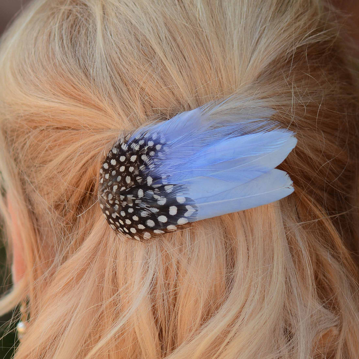 A close-up of a hair accessory clipped into blonde hair, featuring a black and white spotted with light blue feathers.
