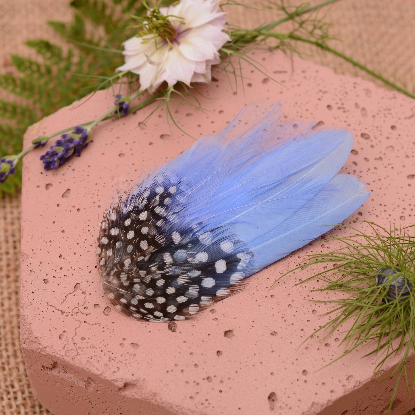 Blue feathered hair clip on a pink surface with flowers and greenery.