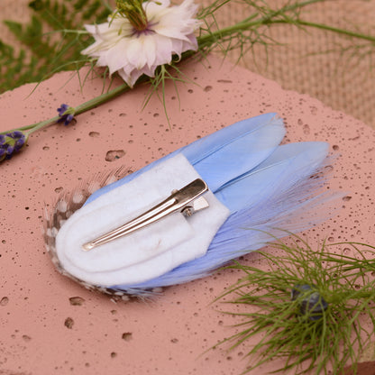 Blue feathered hair clip on a pink surface with flowers and greenery.