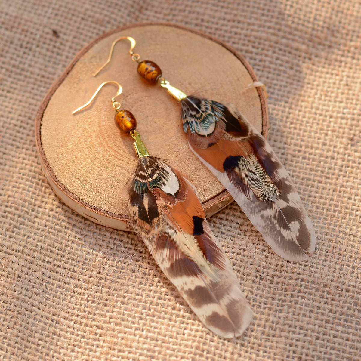 A pair of dangling natural pheasant feather earrings with gold fishhook wires and amber beads, displayed on a round wooden slice over a beige burlap fabric background.