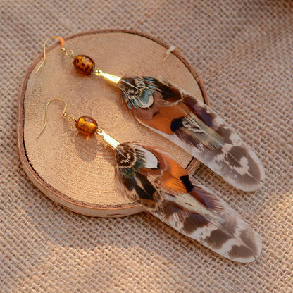 A pair of statement feather drop earrings with amber beads and gold hooks, displayed on a round wooden slice over a textured beige fabric background.