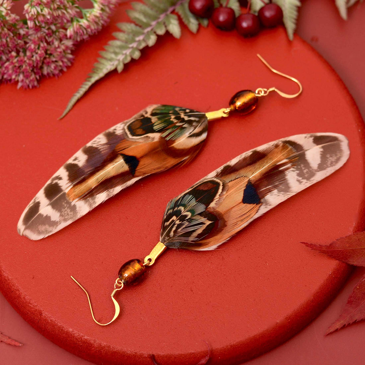 A pair of dangling earrings made from large patterned real feathers with gold hooks and amber-brown beads, arranged on a red circular surface with decorative leaves and berries. Perfect colours for fall and autumn with a bohemian feel.