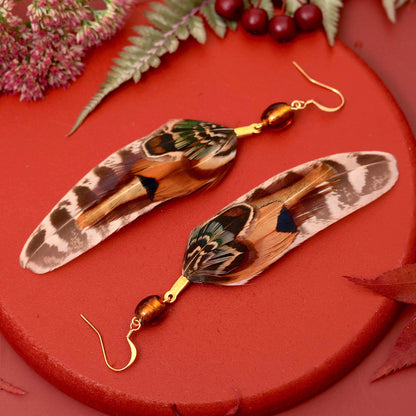 A pair of dangling earrings made from large patterned real feathers with gold hooks and amber-brown beads, arranged on a red circular surface with decorative leaves and berries. Perfect colours for fall and autumn with a bohemian feel.
