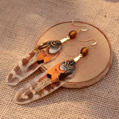 A pair of bohemian feather earrings with gold-tone hooks and amber beads, displayed on a round wooden slice over a burlap fabric background.