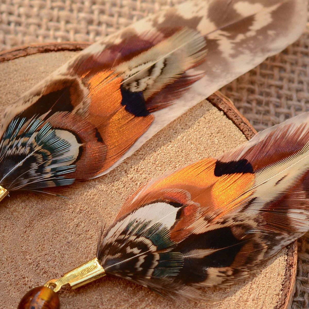 Two decorative feather earrings with intricate multicolor patterns and light reflecting qualities, including orange, brown, black, teal, and cream, attached to gold-toned metal caps and amber beads, displayed on a textured burlap background.