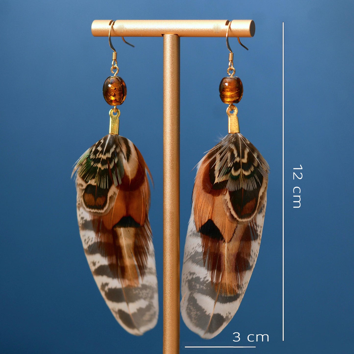 A pair of long feather drop earrings displayed on a gold stand against a blue gradient background, with white measurement lines and text indicating 12 cm length and 3 cm feather width.