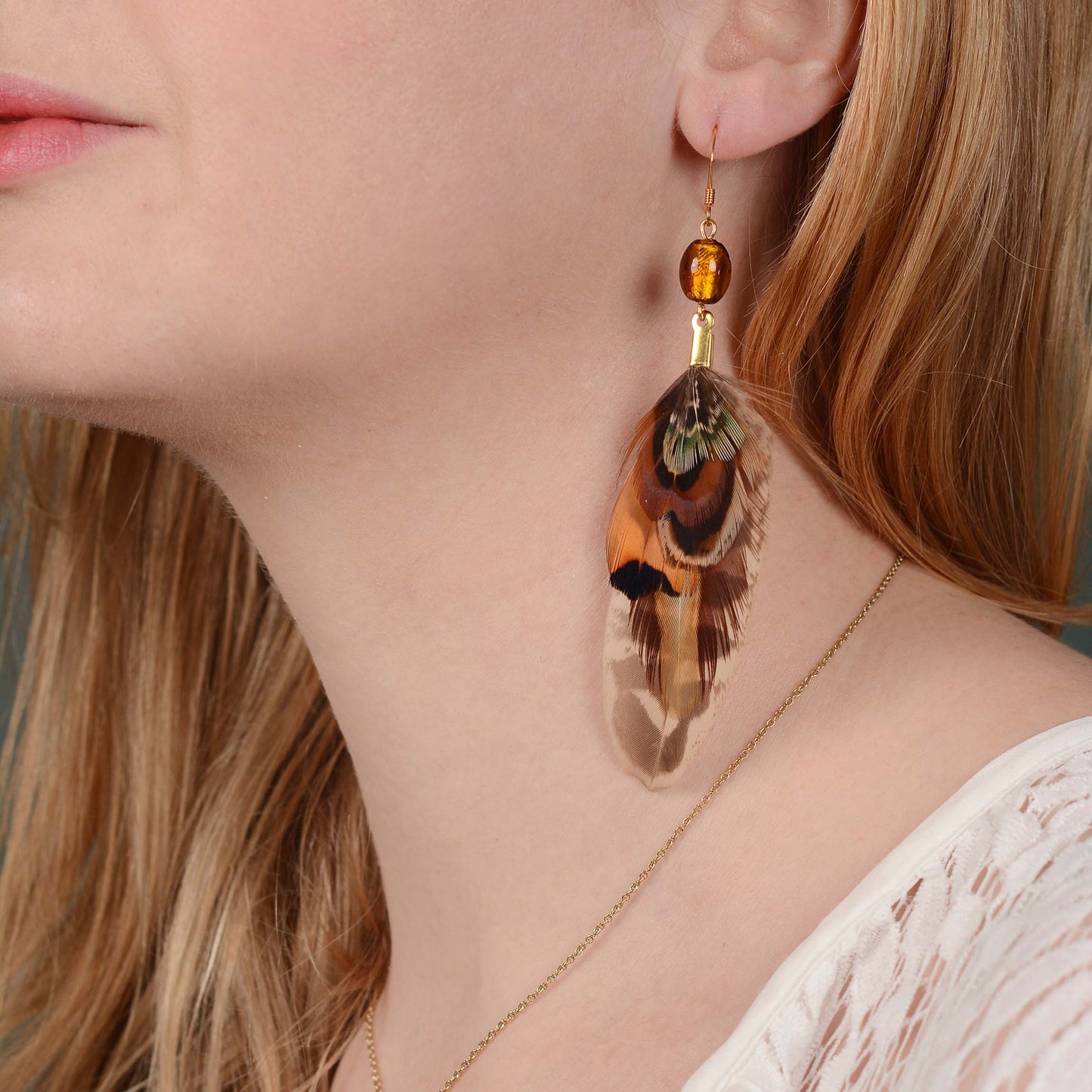 A close-up of a woman wearing a dangling feather earring with a gold-toned hook, amber bead, and a feather featuring brown, orange, black, and green tones. A bohemian style perfect for summer and festivals.