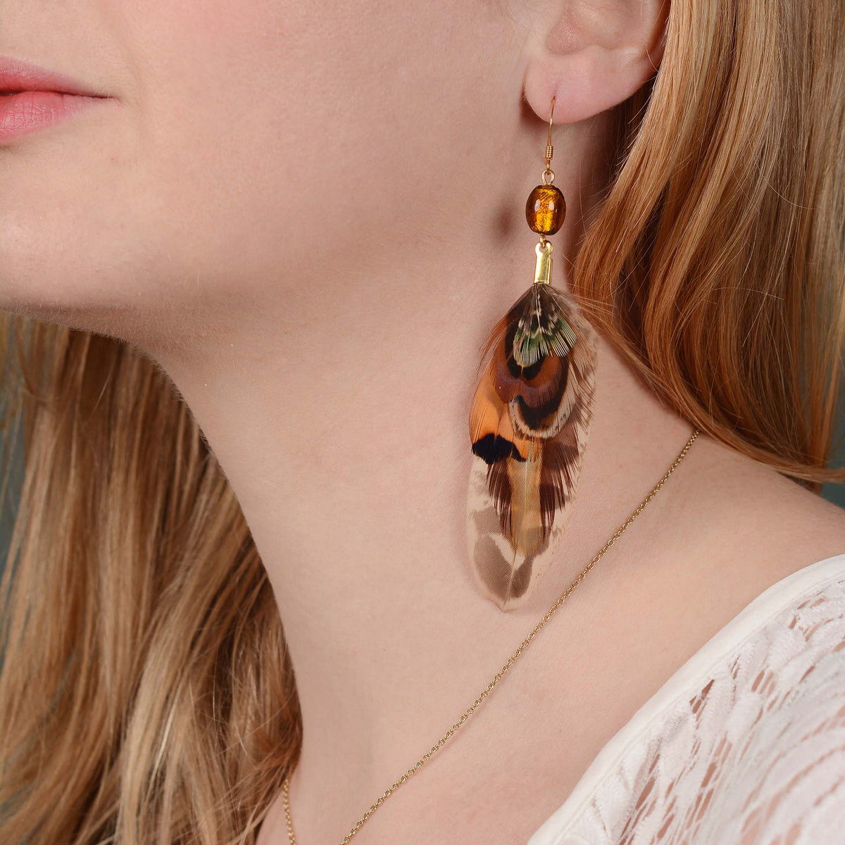 A close-up of a woman wearing a dangling feather earring with a gold-toned hook, amber bead, and a feather featuring brown, orange, black, and green tones. A bohemian style perfect for summer and festivals.