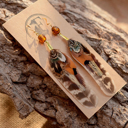 A pair of dangling feather earrings with amber beads and gold hooks, displayed on a kraft card over a textured piece of wood bark.