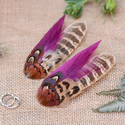 Two pheasant feather hair clip fascinators with vibrant purple and brown patterns on a textured fabric background.