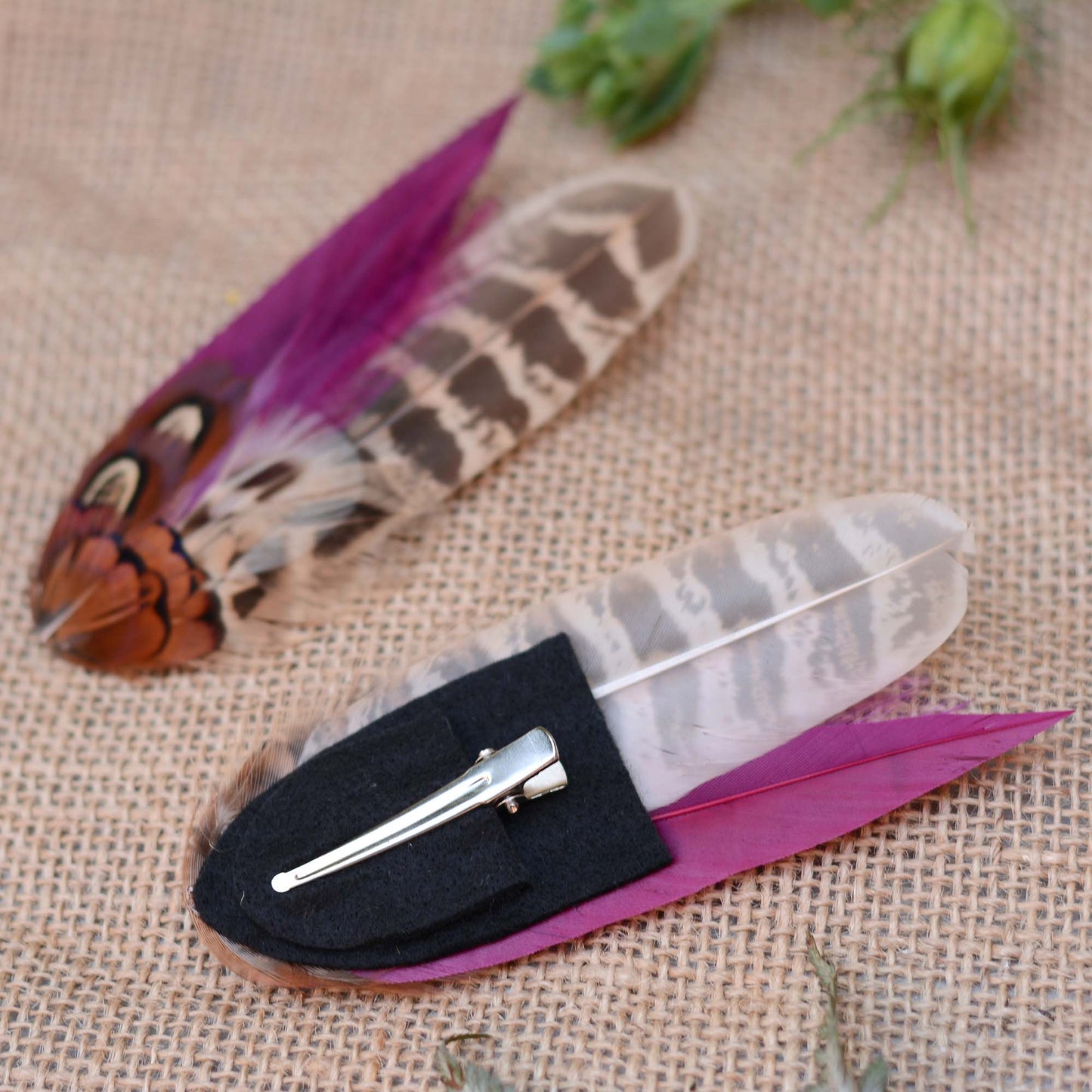 Two pheasant feather hair clip fascinators with vibrant purple and brown patterns on a textured fabric background. One is upside down to show the metal hair clip.