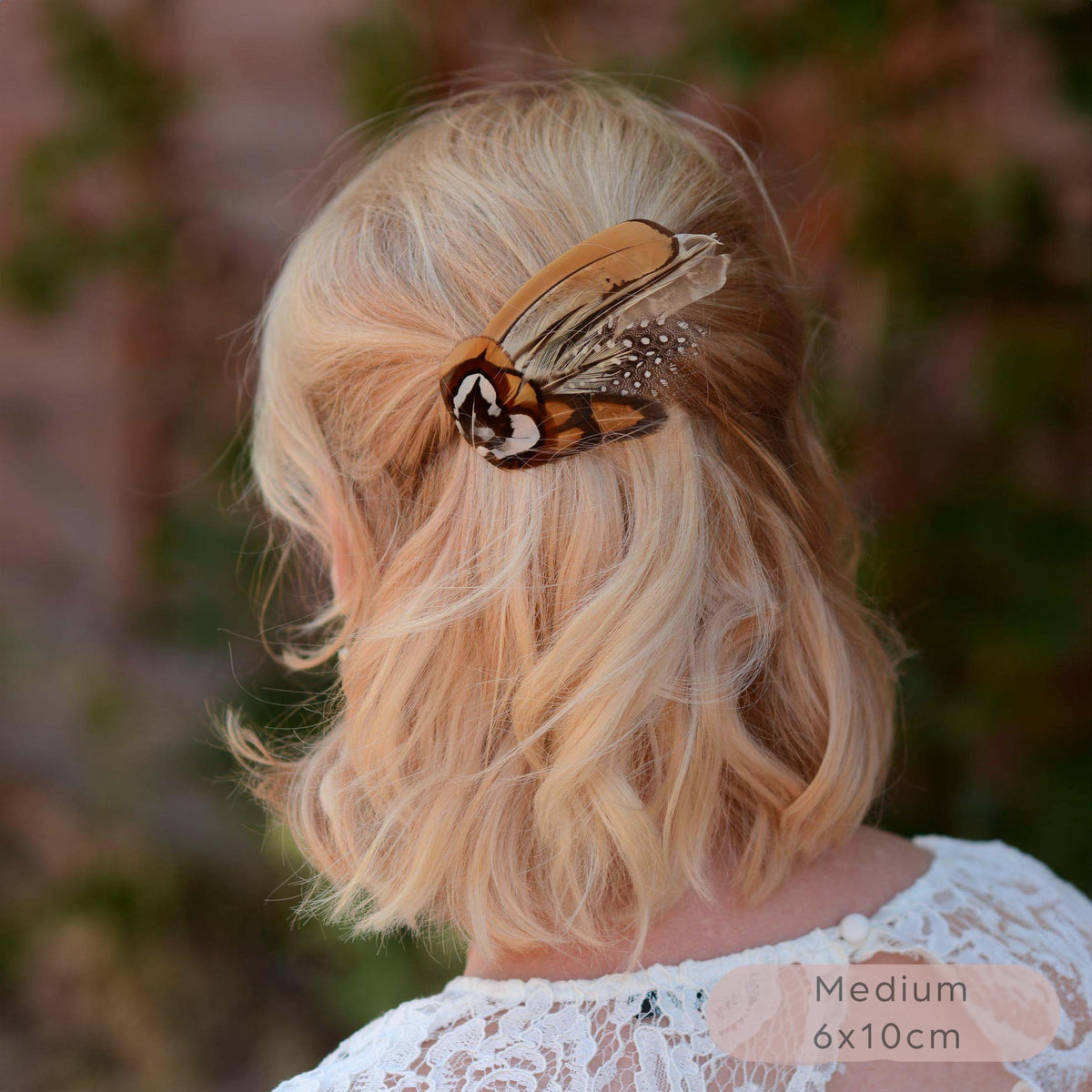 A small  wing-shaped pheasant feather hair clip with a black, brown and white patterned feathers, shown securing a blonde woman's half-up hairstyle outdoors. 