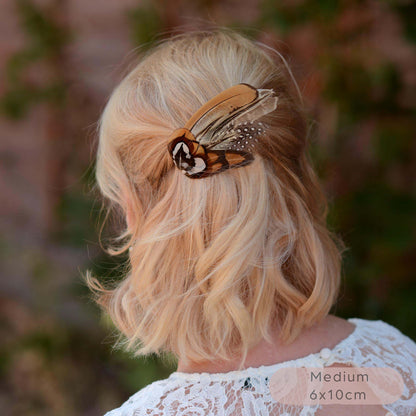A small  wing-shaped pheasant feather hair clip with a black, brown and white patterned feathers, shown securing a blonde woman's half-up hairstyle outdoors. 